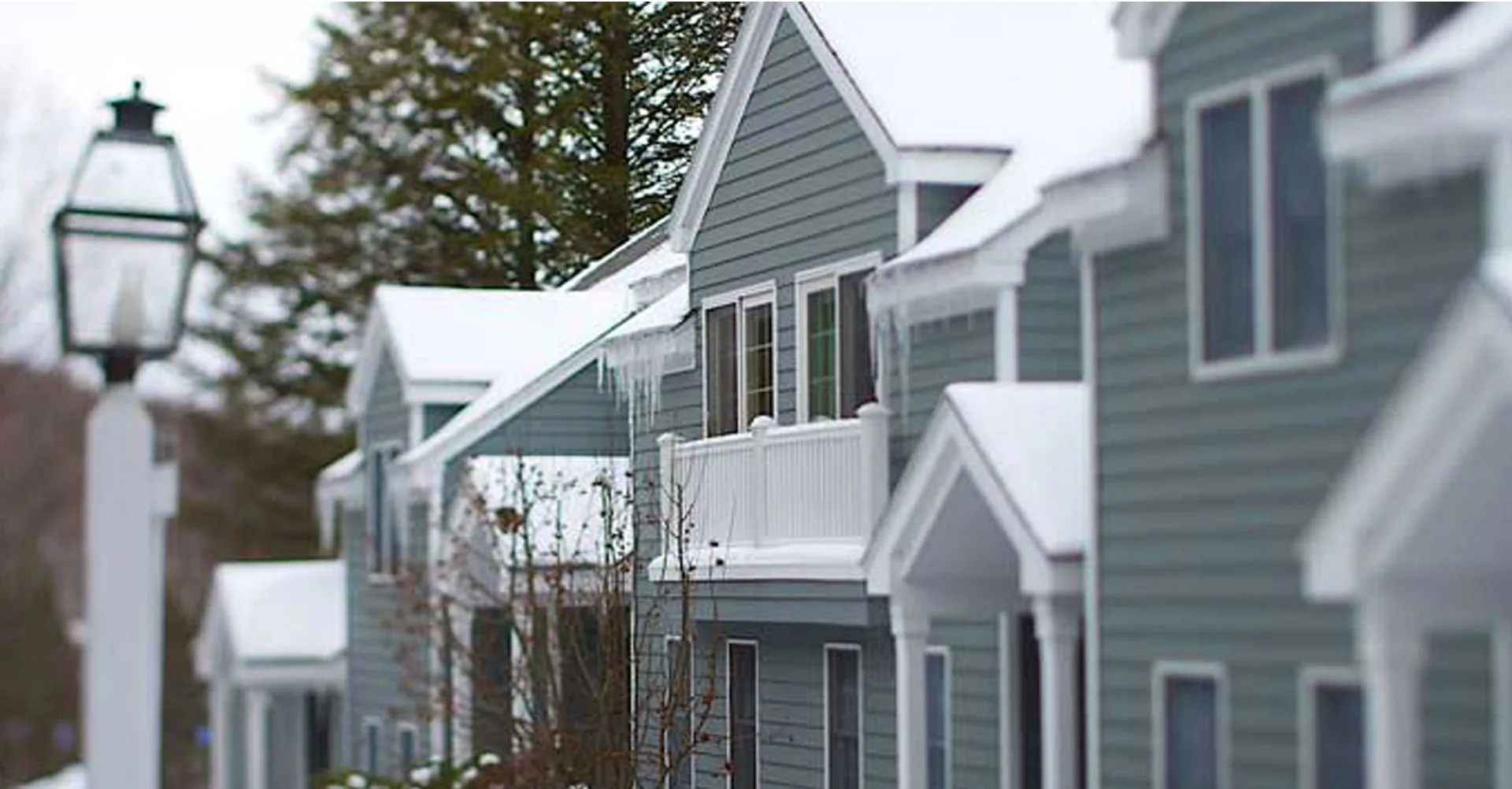 Row of blue-sided townhomes with snow-covered roofs and icicles beside a white lamppost at Jiminy Peak