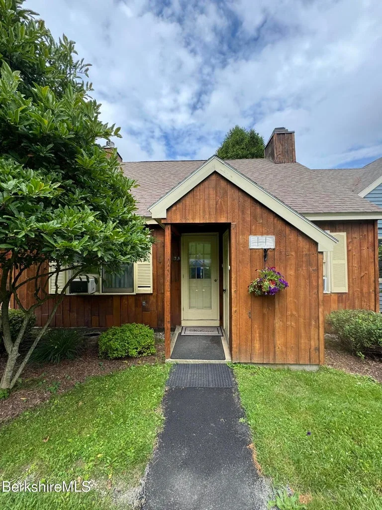 Wood-sided Country Village townhouse entrance with peaked roof, hanging flower basket, and walkway to door