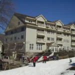 Four-story J.J.'s Lodge condominium building with balconies and skiers gathered on snow at the base